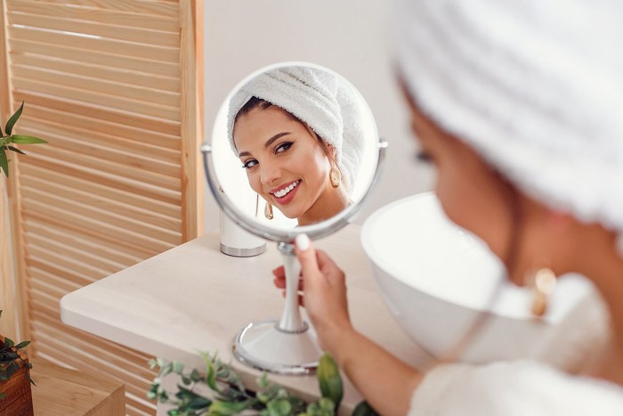 1167975260 Attractive smiling woman with a white towel on her head dressed in bathrobe looks at herself in the mirror in stylish bathroom after morning shower. Beauty concept.