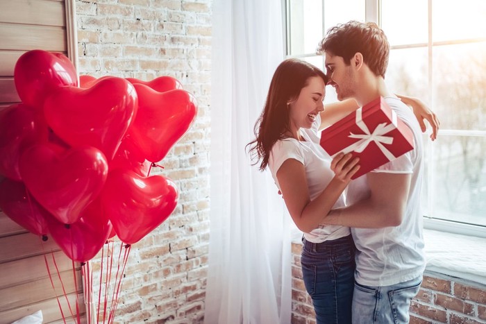 Beautiful young couple at home. Hugging, kissing and enjoying spending time together while celebrating Saint Valentines Day with gift box in hand and air balloons in shape of heart on the background.