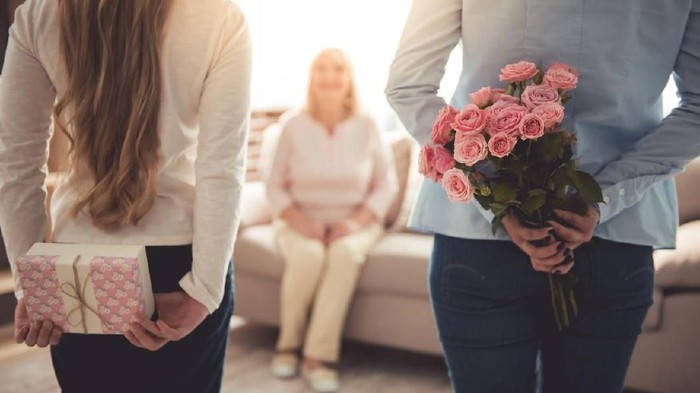Teenage girl and her mom are hiding flowers and a gift box for their beautiful granny behind backs while grandma is sitting on couch at home
