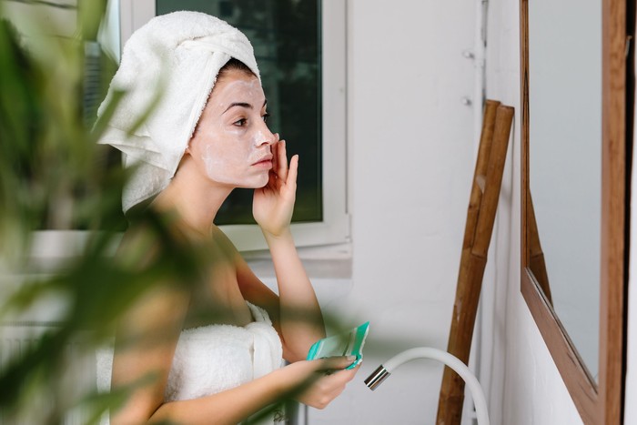 beautiful young woman with towel wrapped around her head is applying mask on her face in front of the mirror at home. Skin care.