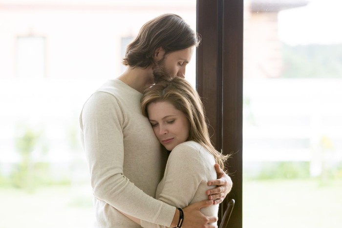 zodiak Sad young married couple embracing standing in living room opposite window at home. Sorrowful wife and husband feels unhappy, thinking about problems in relations, miscarriage, misbirth or infertility