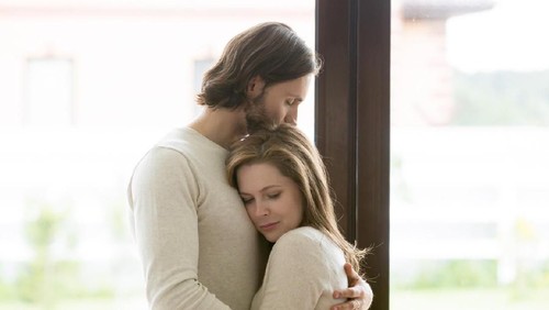 Sad young married couple embracing standing in living room opposite window at home. Sorrowful wife and husband feels unhappy, thinking about problems in relations, miscarriage, misbirth or infertility
