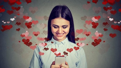 Beautiful happy woman sending love text message on mobile phone with red hearts flying away from screen isolated on gray background. Positive human emotions face expressions