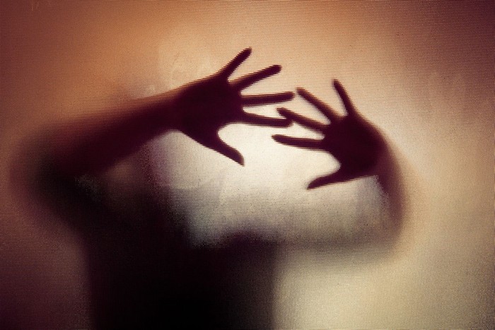 Colour backlit image of the silhouette of a woman with her hands pressed against a glass window. The silhouette is distorted, and the arms elongated, giving an alien-like quality. The image is sinister and foreboding, with an element of horror. It is as if the woman is trying to escape from behind the glass. Horizontal image with copy space.