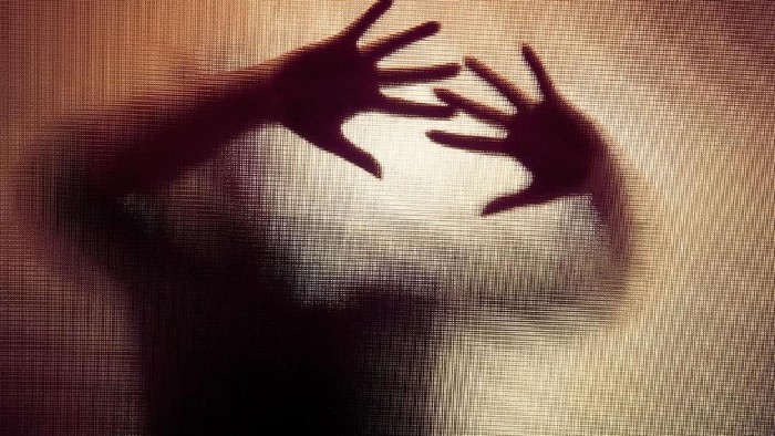 Colour backlit image of the silhouette of a woman with her hands pressed against a glass window. The silhouette is distorted, and the arms elongated, giving an alien-like quality. The image is sinister and foreboding, with an element of horror. It is as if the woman is trying to escape from behind the glass. Horizontal image with copy space.