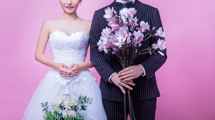 Portrait of multi-ethnic wedding couple holding flowers while standing against pink background