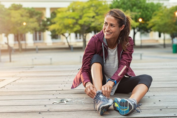 1040307846 Mature fitness woman tie shoelaces on road. Cheerful runner sitting on floor on city streets with mobile and earphones wearing sport shoes. Active latin woman tying shoe lace before running.