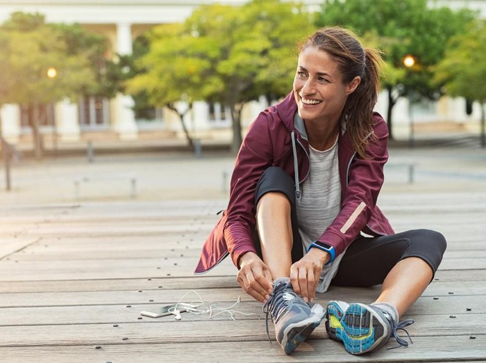 Mature fitness woman tie shoelaces on road. Cheerful runner sitting on floor on city streets with mobile and earphones wearing sport shoes. Active latin woman tying shoe lace before running.