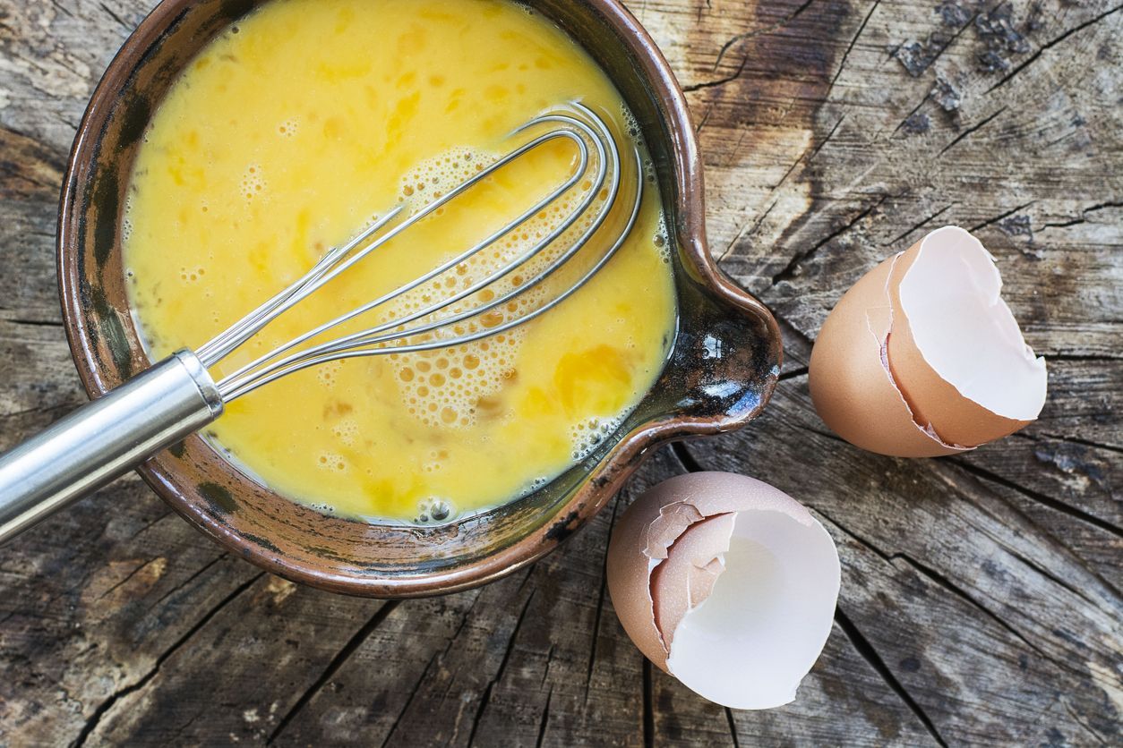 telur dadar Beaten eggs and whisk in a ceramic bowl. In this photograph the bowl of eggs is placed on a rustic wooden surface, the whisk is placed in the bowl of eggs and is projecting out of the photograph. The egg shells from the beaten eggs are placed on the wooden surface. This photograph is taken with natural light. The image is intended for chefs, cooks, restaurants and egg sellers, as well as other.