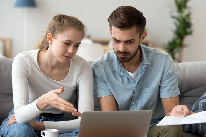 zodiak Focused man and woman sit on couch using laptop taking care of financial bills, husband and wife manage bank documents, reading regulations online, upset couple consider received papers