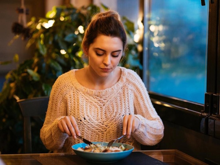Beautiful girl having supper at restaurant, using cutlery for eating salad. Dressed in knitted sweater.