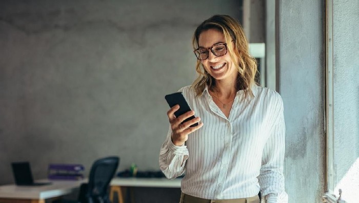 Smiling businesswoman using phone in office. Small business entrepreneur looking at her mobile phone and smiling.