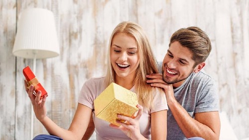 What a wonderful gift. Surprised young woman is opening box and smiling. Man is sitting and embracing her with love