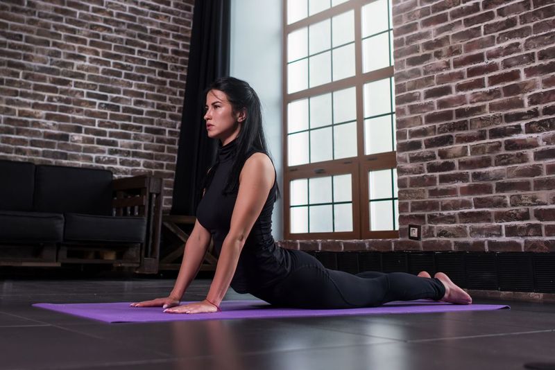 Female yoga beginner doing bhujangasana cobra pose on mat in loft apartment.