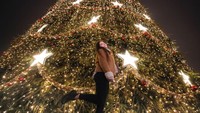 Seorang warga di Romania terlihat asyik berpose di depan pohon Natal yang didekorasi penuh dengan lampu kelap-kelip. (AP Photo/Vadim Ghirda)