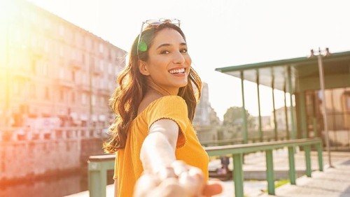 Happy casual girl holding hand and looking behind while walking in the city street. Beautiful latin woman walking around the city on a sunny day. Young smiling woman pulling her boyfriend and looking at camera.