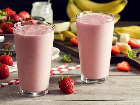 Two cold strawberry banana yogurt smoothies in pint glasses with striped straws. The berries and bananas are in the background in a colander and on a cutting board. A jar of yogurt is also in the background. Everything is on a faded wood picnic table.