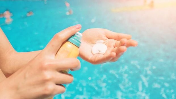 Suntan lotion. Woman applying sunscreen solar cream on face. Beautiful happy cute girl puts suntan cream from plastic container bottle on her nose and cheecks. Skincare concept. Female in straw hat and bikini on beach.