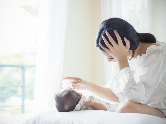 Asian Mother nursery feeding bottle of formula milk to newborn baby in bed suffering from post natal depression. Health care single mom motherhood stressful concept.