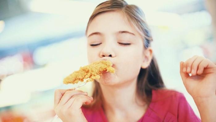 Closeup front view of cute little girl eating fried chicken and yes,enjoying it. She has a piece of chicken stuck on a fork and biting it with pleasure. Her eyes are closed, the girl is wearing pink t-shirt.