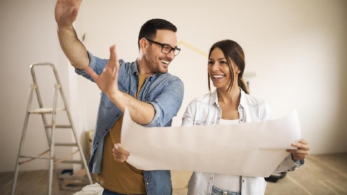 Young couple standing in their apartment while woman holding blueprint and man showing to her new ideas.