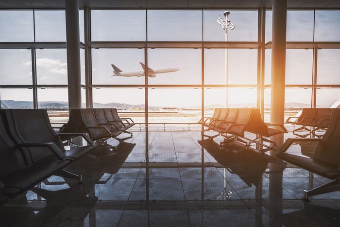 Wide-angle view of a modern aircraft gaining the altitude outside the glass window facade of a contemporary waiting hall with multiple rows of seats and reflections indoors of an airport terminal El Prat in Barcelona
