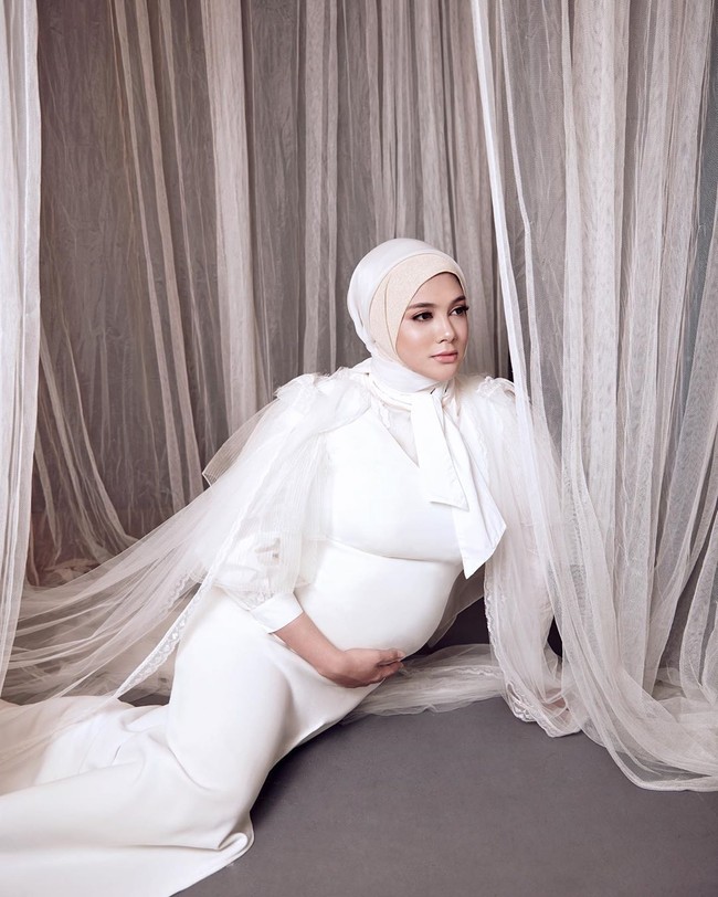 Foto kehamilan selanjutnya, Biby yang mengenakan dress putih berpose duduk sambil menyentuh perutnya. Wanita 29 tahun itu menggunakan hijab dua layer dan diikat pita di bagian bawah lehernya. Foto: instagram @bibyalraen13