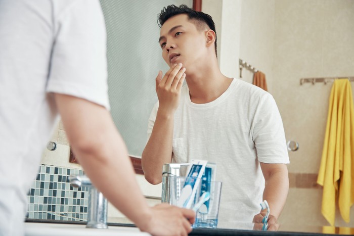 From below back view of man touching face skin looking at reflection of bathroom mirror at home