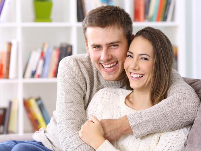 Front view portrait of a happy couple with perfect smile looking at you sititng on a couch in the living room at home in winter