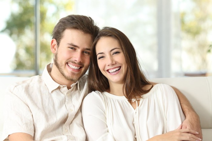 Front view portrait of a happy couple with perfect smile looking at you sititng on a couch in the living room at home in winter