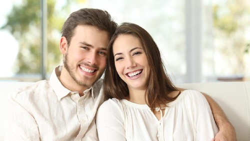 Front view portrait of a happy couple with perfect smile looking at you sititng on a couch in the living room at home in winter