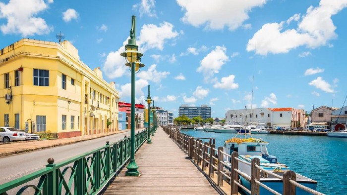 Bright image of wooden promenade at the waterfront of Bridgetown in Barbados. Colorful building against blue sky with white clouds. Boats and yachts in the harbor.