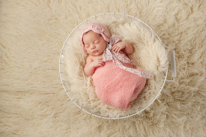 A portrait of a five week old newborn baby girl wearing a pink bonnet and sleeping in a wire basket. Shot from overhead.