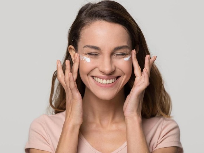 Smiling happy pretty young teen girl woman applying moisturizing lotion cosmetic cream on face for clean healthy soft skin care natural beauty treatment isolated on white grey studio blank background