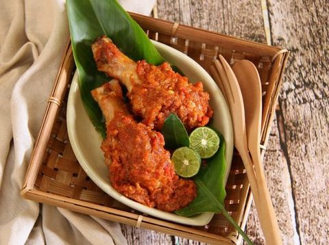 Ayam Rica-Rica, the popular and traditional dish from Manado, chicken braised in spicy red chili paste. Two drumsticks are arranged on a small ceramic plate that has been lined with a banana leaf. Two lime halves and a lime leaf are used to garnish the dish. The plate is placed on a square bamboo plate with a pair of wooden cutlery tucked in. The bamboo plate is placed on a rustic wooden table.
