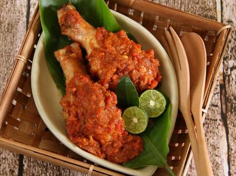 Ayam Rica-Rica, the popular and traditional dish from Manado, chicken braised in spicy red chili paste. Two drumsticks are arranged on a small ceramic plate that has been lined with a banana leaf. Two lime halves and a lime leaf are used to garnish the dish. The plate is placed on a square bamboo plate with a pair of wooden cutlery tucked in. The bamboo plate is placed on a rustic wooden table.