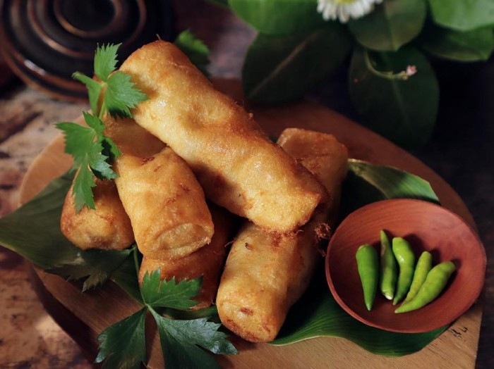 Sosis Solo, the fried egg roll with minced beef stuffing from Solo (Surakarta), Central Java. The egg rolls are plated on a wooden cutting board lined with banana leaf and garnished with celery sprigs. Accompanied with green chili peppers.