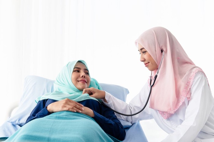 hijab dokter A young beautiful Muslim doctor is using a stethoscope to listen to the patients heartbeat at the bed in the hospital examination room.