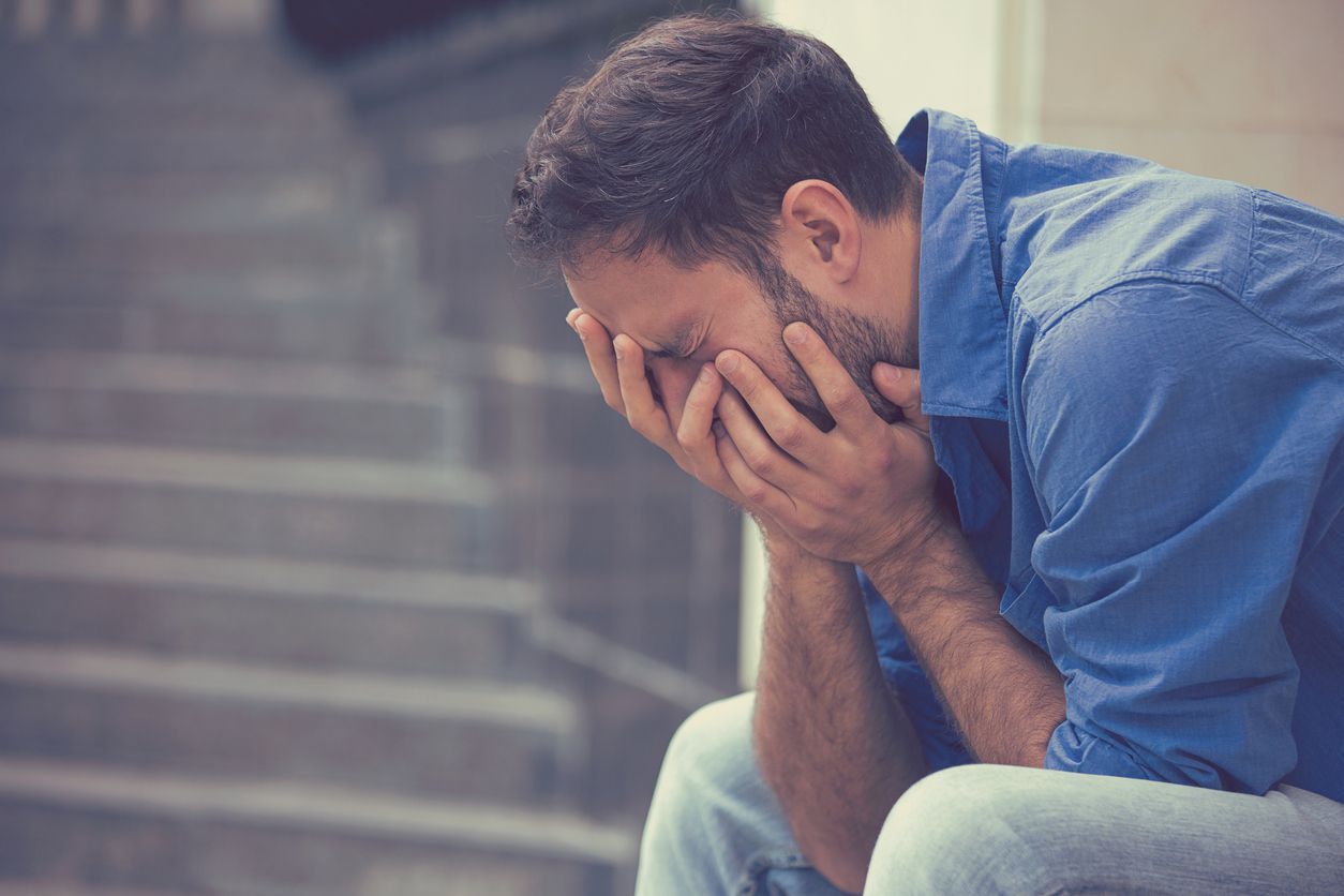 side profile stressed sad young crying man sitting outside holding head with hands looking down. Human emotion feelings