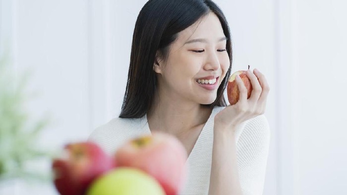 Makan buah healthy beautiful attractive long black hair woman happiness smile hand hold red apple with foreground of fruit bowl white room interior background