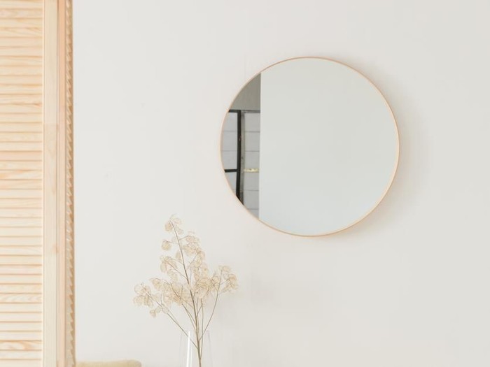 Round mirror hanging on white wall in real photo of bright sitting room interior with wooden cupboard with books and plant in glass vase