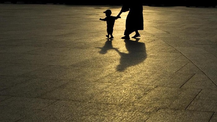A young child and an adult walking across pavement at night while holding hands.  The silhouette and shadow of the two in the dark can be seen.