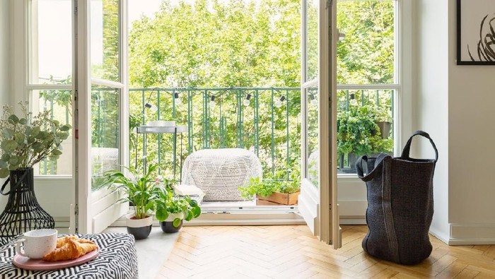 Open glass door from a living room interior into a city garden on a sunny balcony with green plants and comfy furniture