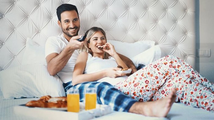 Closeup front view of a mid 20s happy couple watching tv in bed on lazy weekend morning. They are eating some snacks and having sodas. The guy is changing the channels.