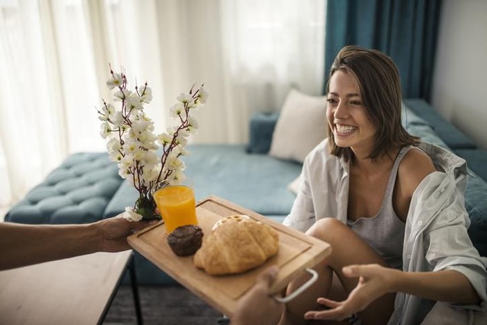 Closeup front view of a mid 20s happy couple watching tv in bed on lazy weekend morning. They are eating some snacks and having sodas. The guy is changing the channels.
