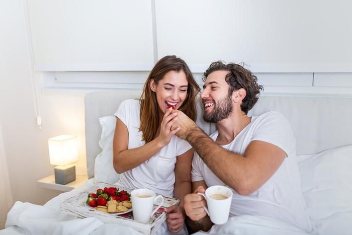 Closeup front view of a mid 20s happy couple watching tv in bed on lazy weekend morning. They are eating some snacks and having sodas. The guy is changing the channels.