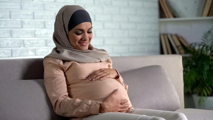 Expecting muslim woman stroking belly sitting on sofa, carrying child harmony