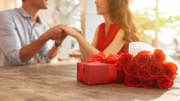 Cheerful young couple sitting at wooden table of outdoor cafe and holding hands while celebrating Valentines Day, gift box and bouquet of red roses on foreground