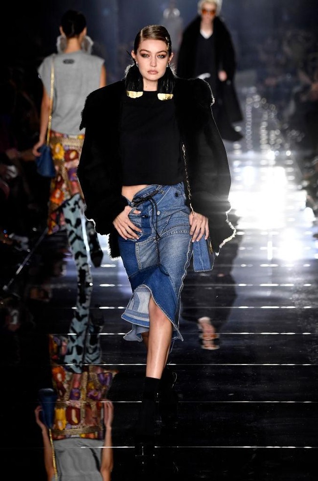 Pada penampilan kedua untuk fashion show Tom Ford, Gigi Hadid tampil edgy dengan rok denim dan coat hitam yang memiliki detail emas di bagian kerah. Foto: Getty Images