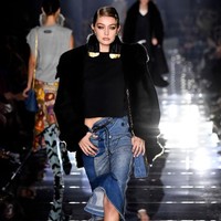 Pada penampilan kedua untuk fashion show Tom Ford, Gigi Hadid tampil edgy dengan rok denim dan coat hitam yang memiliki detail emas di bagian kerah. Foto: Getty Images